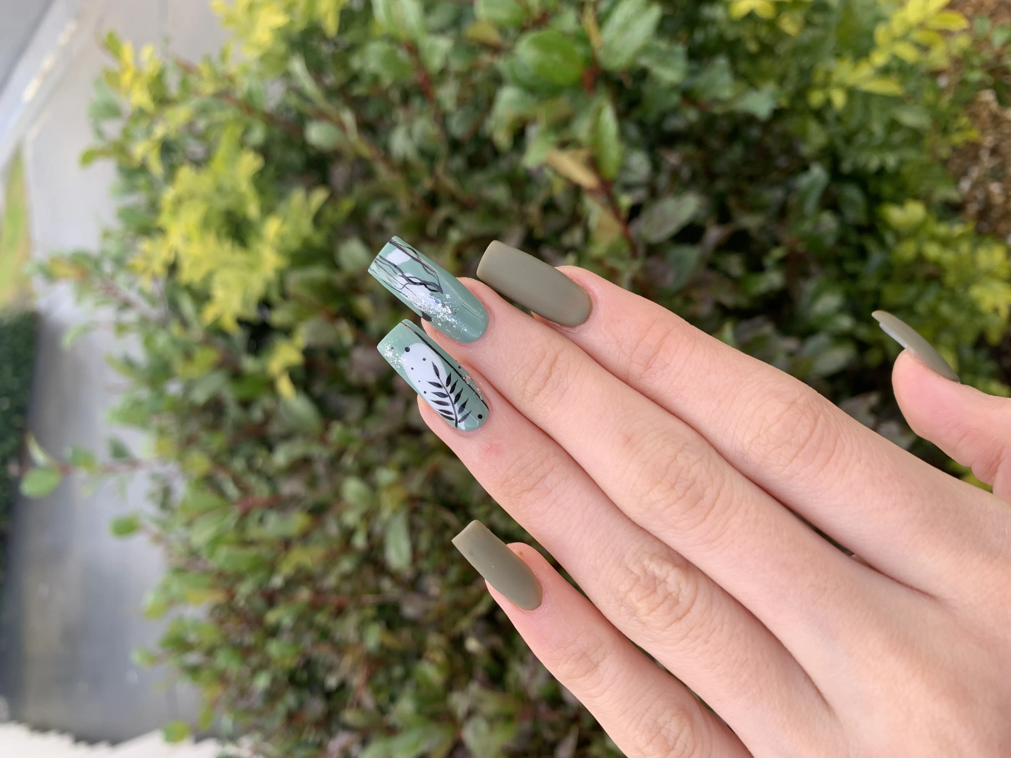Sage-green and matte-olive long coffin nails with black botanical leaf art, outdoors by a hedge